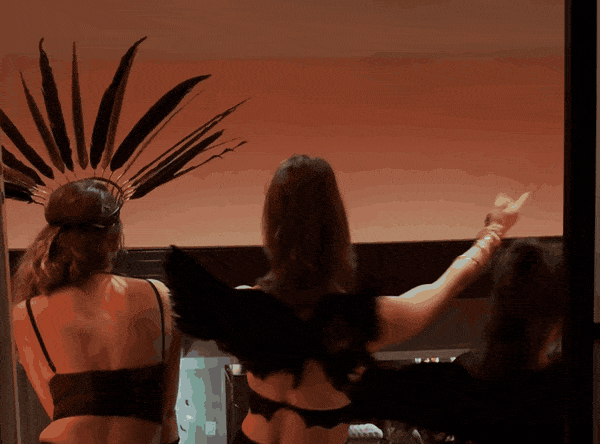 Women in black evening wear and feathers dancing with raised arms against a warm sky